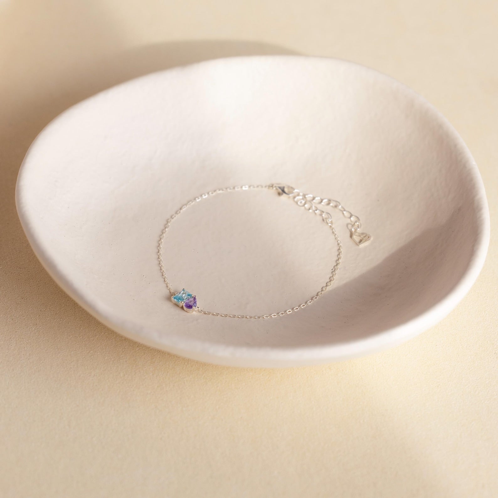 The Toi et Moi Birthstone Bracelet, featuring customizable blue and purple gemstones in a delicate silver design, is displayed in a white ceramic dish.