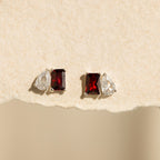Two pairs of Toi et Moi Birthstone Earrings with red rectangular and clear teardrop stones displayed on a textured beige background.