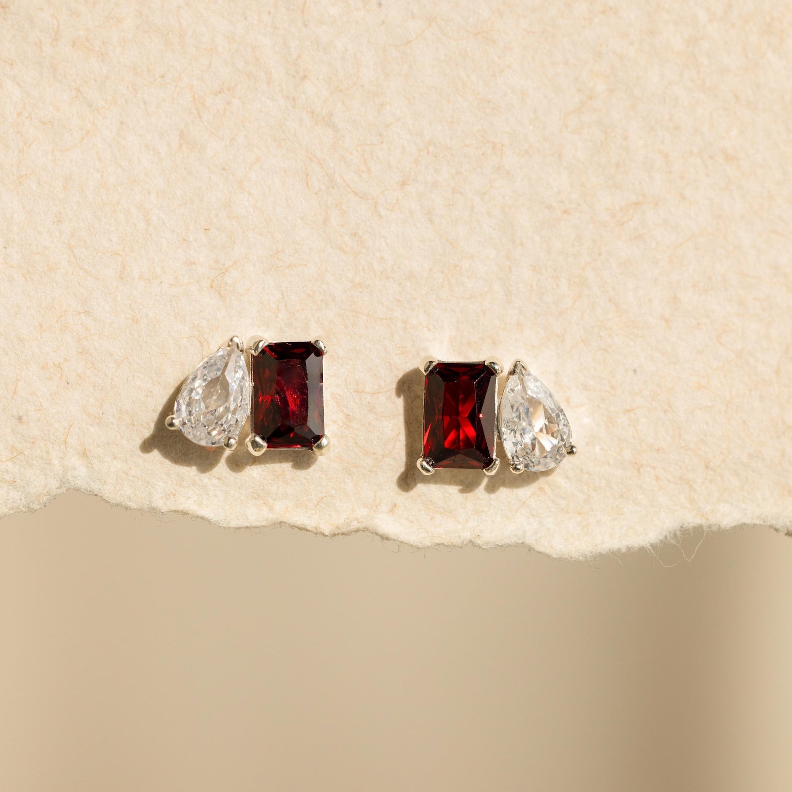 Two pairs of Toi et Moi Birthstone Earrings with red rectangular and clear teardrop stones displayed on a textured beige background.
