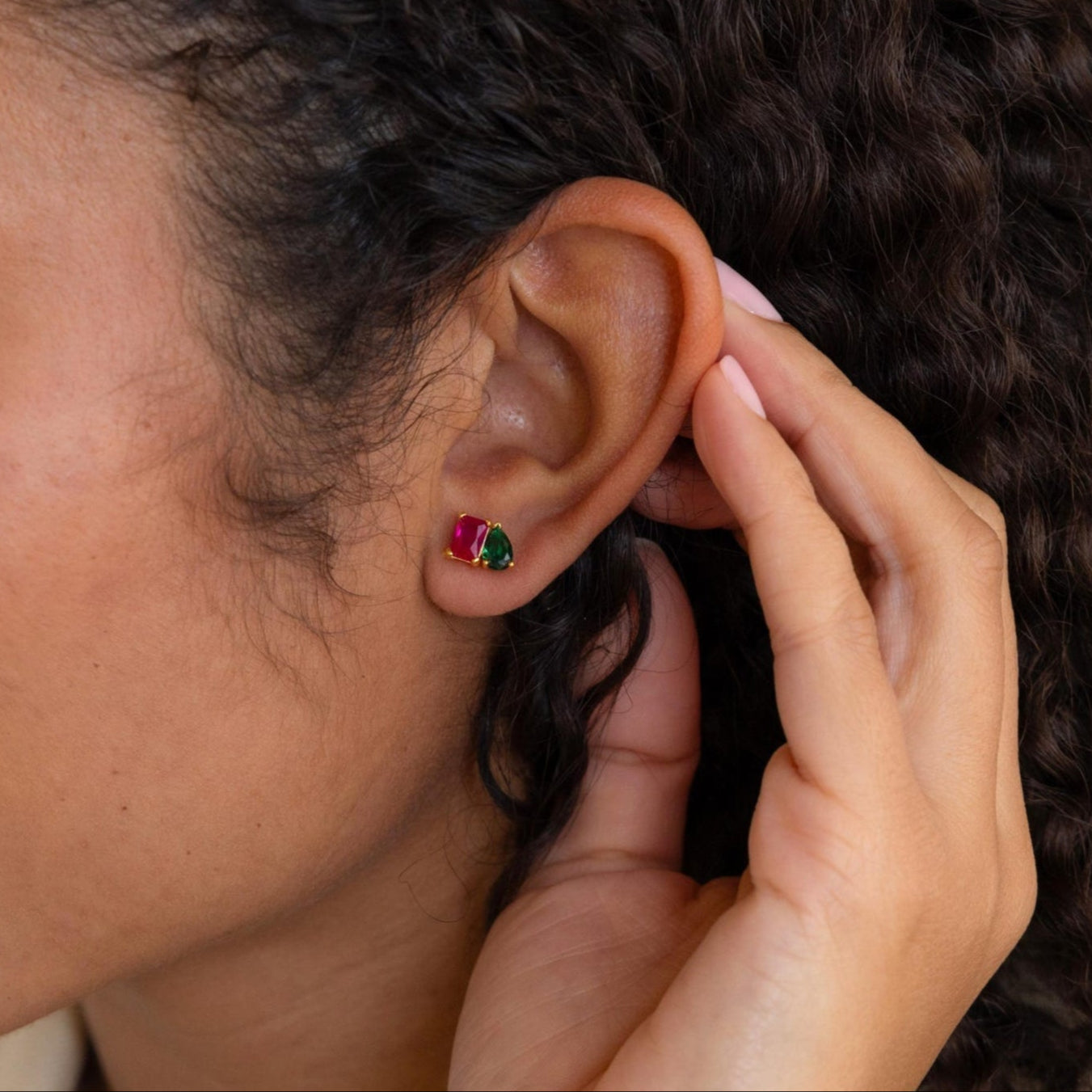 A person with curly hair gently touches their ear while wearing Toi et Moi Birthstone Earrings featuring vibrant red and green stones.