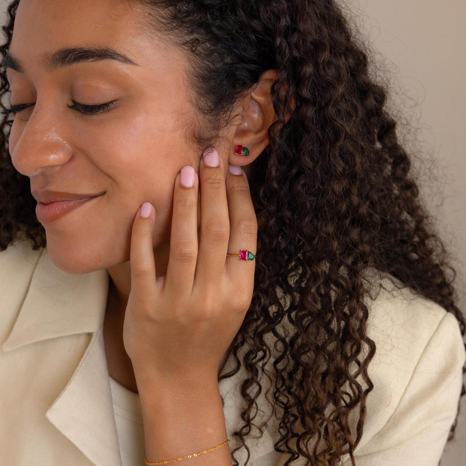 A woman with curly hair smiles, wearing vibrant Toi et Moi Birthstone Earrings and a matching gemstone ring.