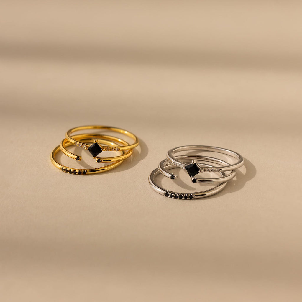 The Black Abyss Ring Set features two stacked sets—one gold, one silver—with dainty geometric bands and black gemstone accents, displayed on a beige background.
