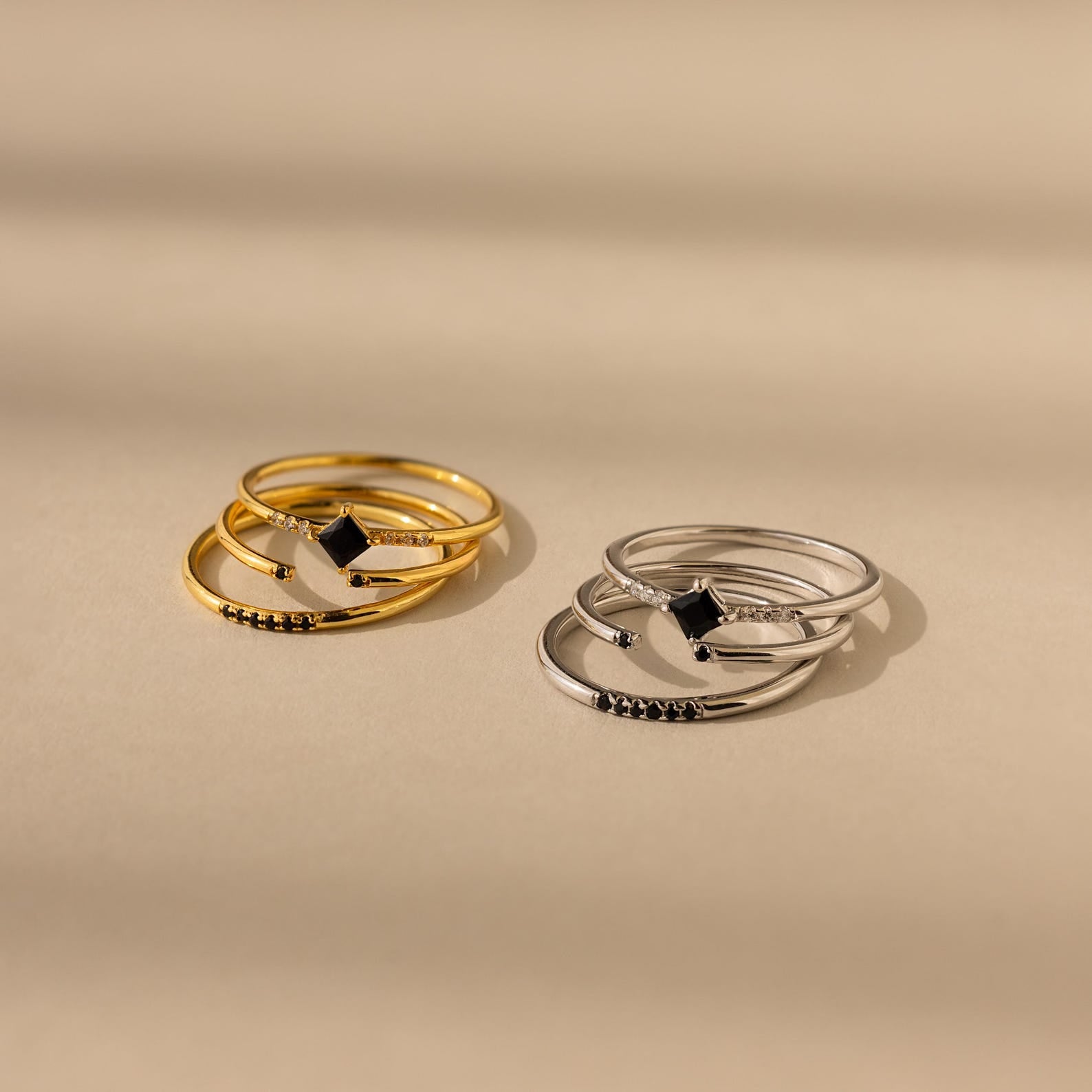 The Black Abyss Ring Set features two stacked sets—one gold, one silver—with dainty geometric bands and black gemstone accents, displayed on a beige background.