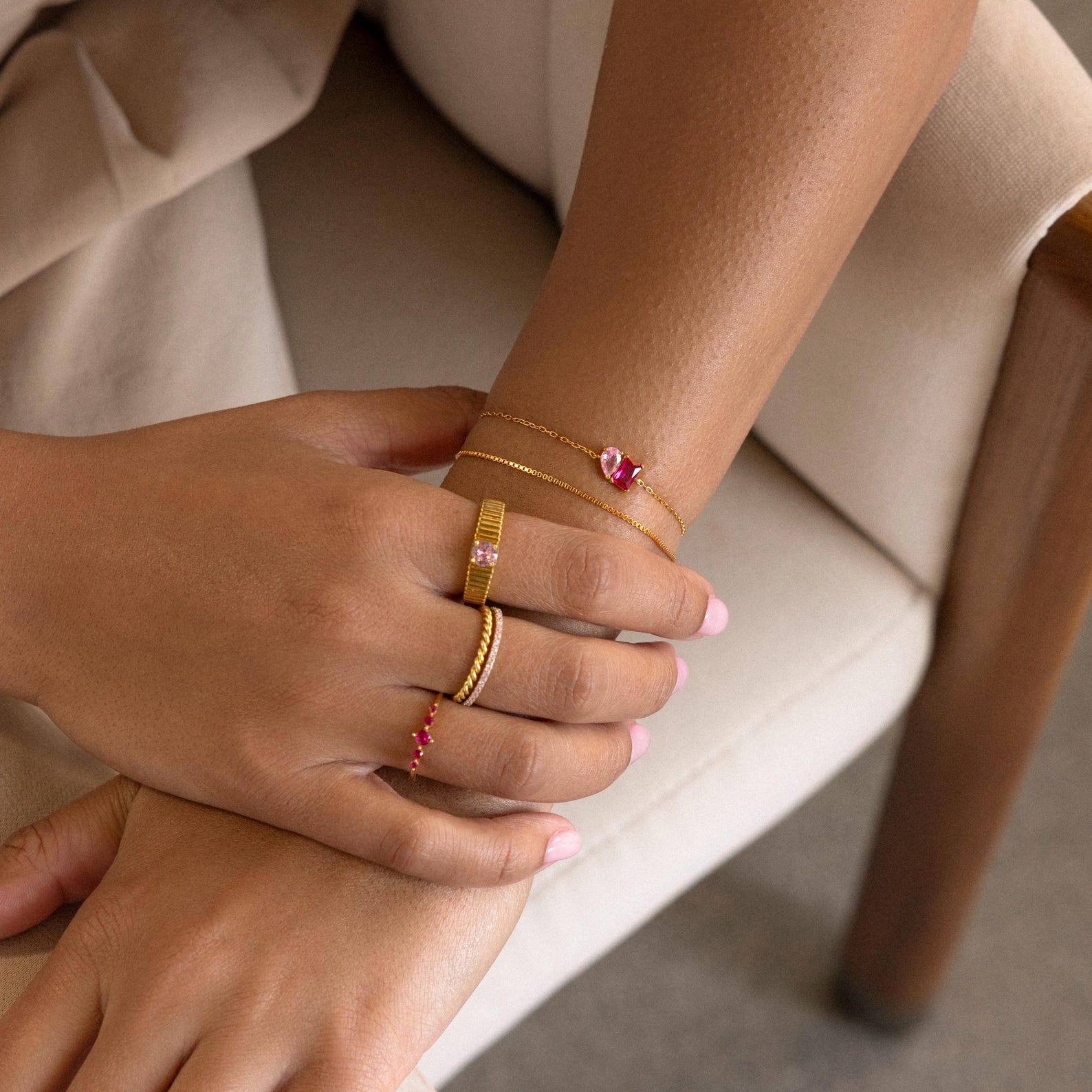 Hands adorned with gold rings and the Toi et Moi Birthstone Bracelet featuring customizable gemstones, resting on a cream chair.