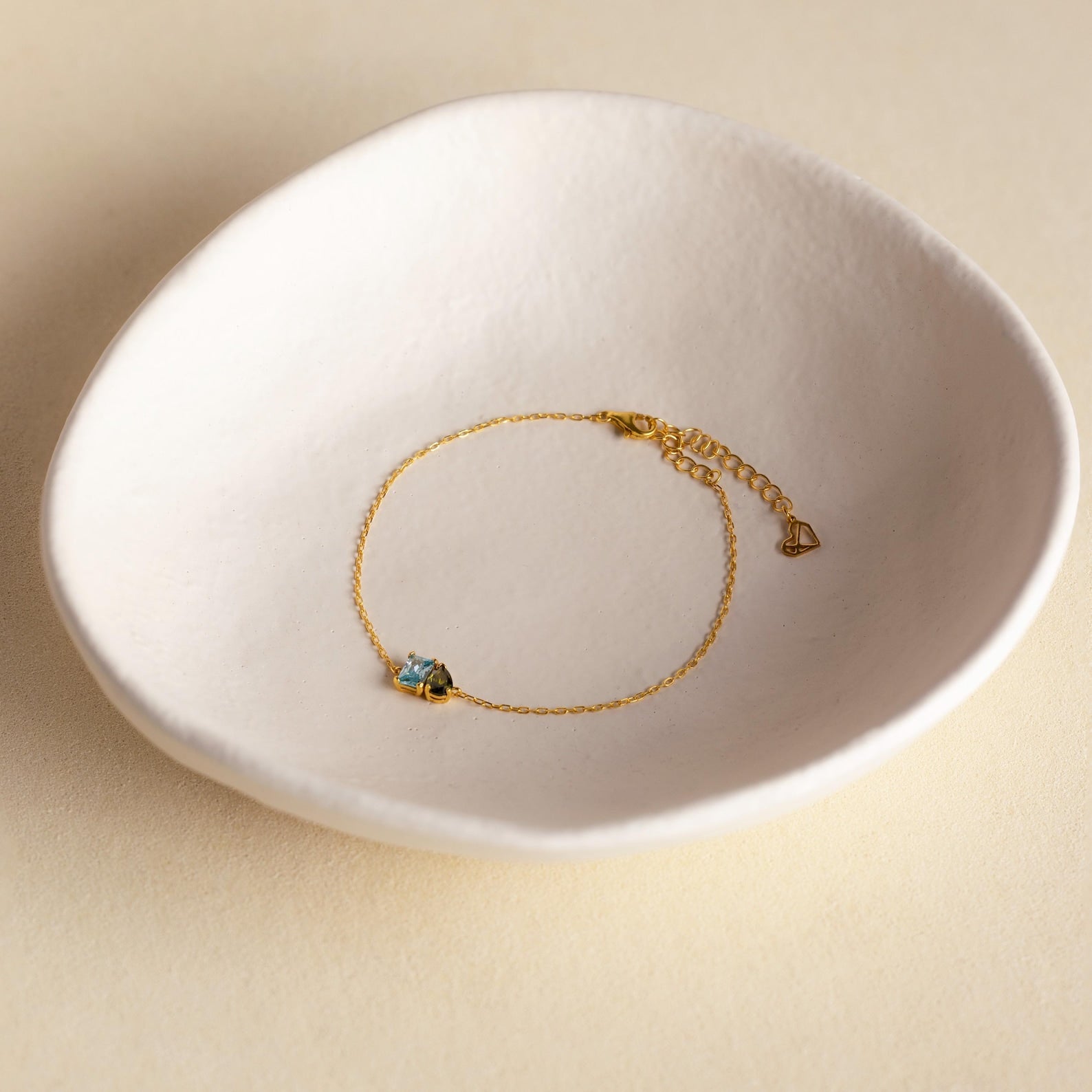 The Toi et Moi Birthstone Bracelet features a delicate gold chain with a small blue gemstone, showcased in a white ceramic dish.