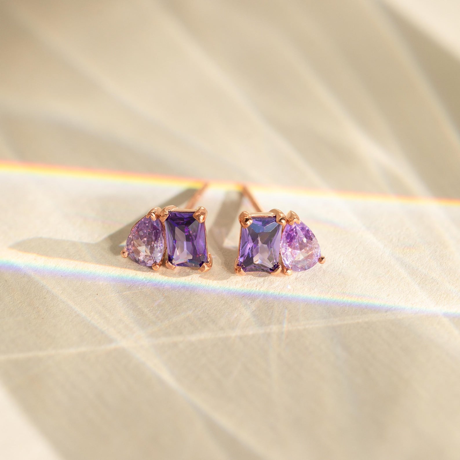 Toi et Moi Birthstone Earrings feature gold studs with rectangular and teardrop purple gemstones, displayed on a light textured surface with rainbow highlights. These earrings make a unique birthstone gift.
