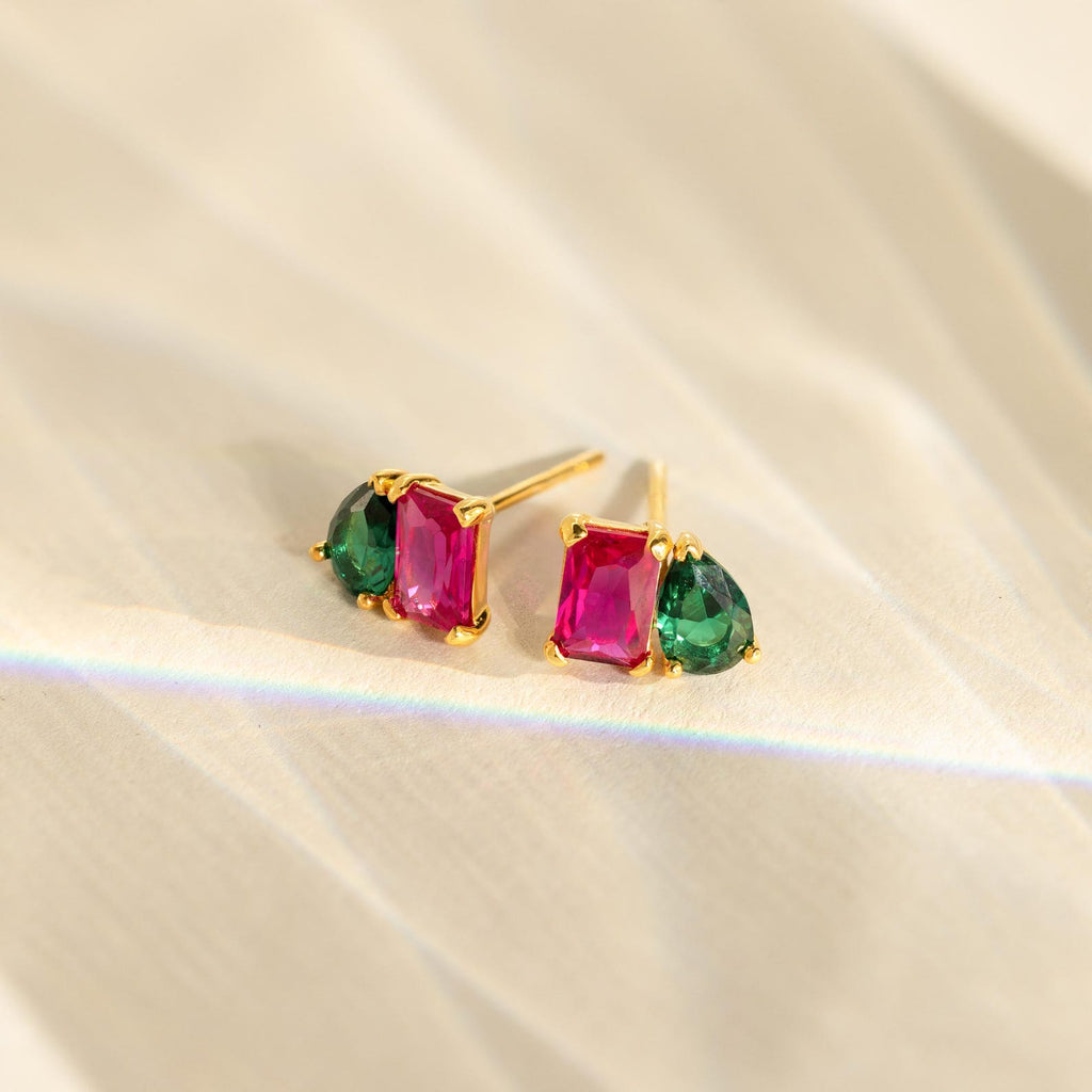 The Toi et Moi Birthstone Earrings feature rectangular pink and teardrop green stones set in gold, elegantly displayed on a textured surface. These vibrant earrings add a touch of color to any outfit.