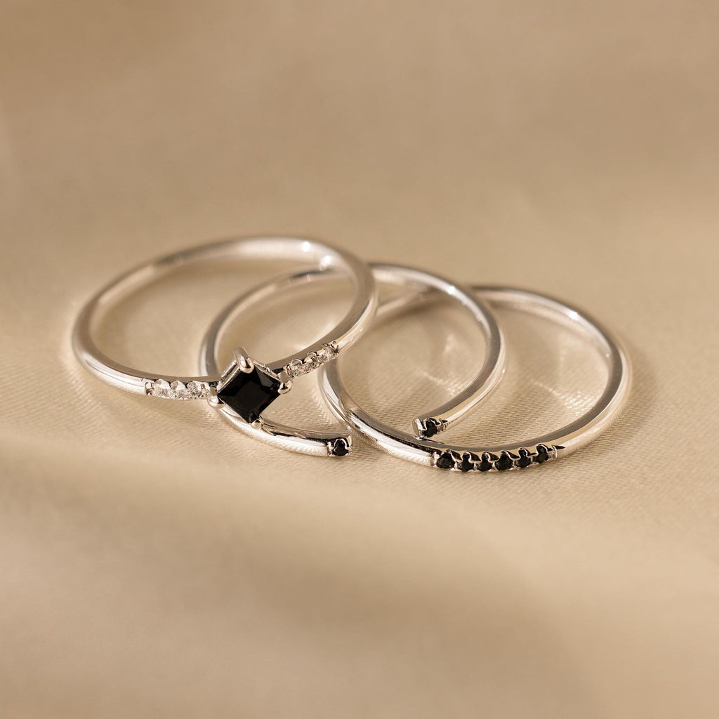 The Black Abyss Ring Set features three delicate silver bands with small black gemstones and white stones, shown on a soft beige fabric background.