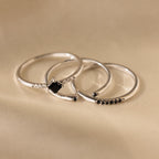 The Black Abyss Ring Set features three delicate silver bands with small black gemstones and white stones, shown on a soft beige fabric background.