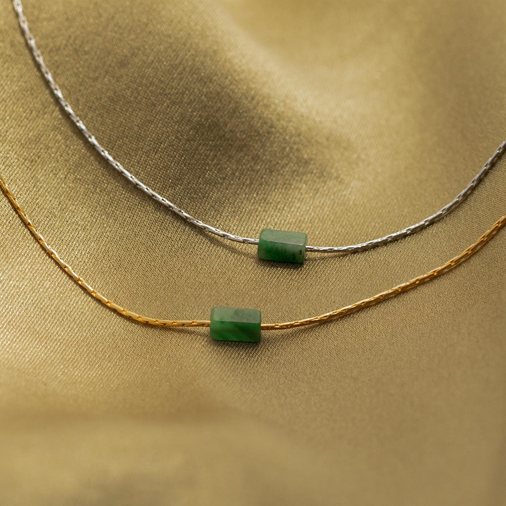 The Jade Bar Jewelry Set in Sterling Silver features two necklaces—one silver, one gold—each with a green rectangular jade bar pendant, displayed on gold fabric. This set offers elegant jade bar jewelry for any occasion.