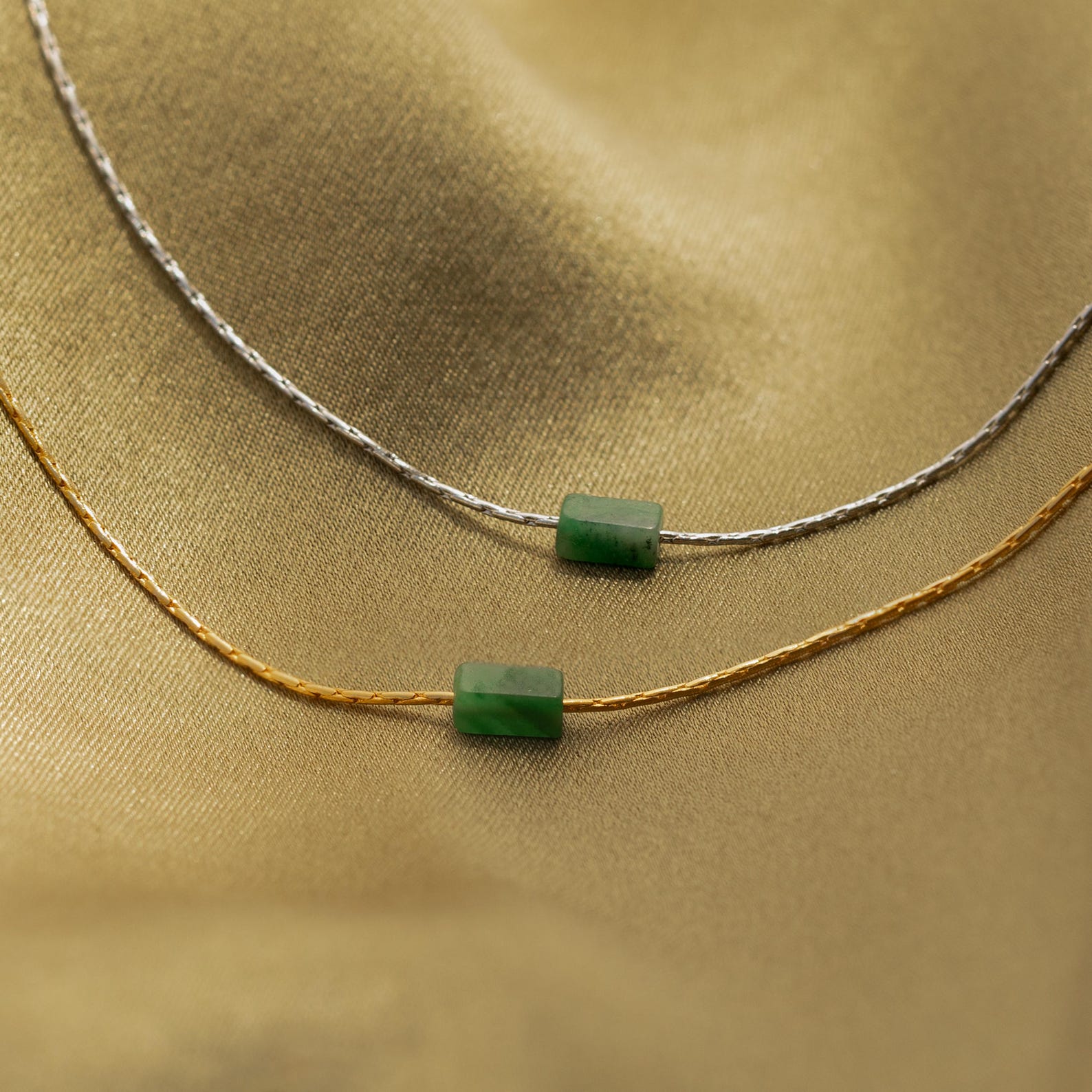The Jade Bar Jewelry Set in Sterling Silver features two necklaces—one silver, one gold—each with a green rectangular jade bar pendant, displayed on gold fabric. This set offers elegant jade bar jewelry for any occasion.