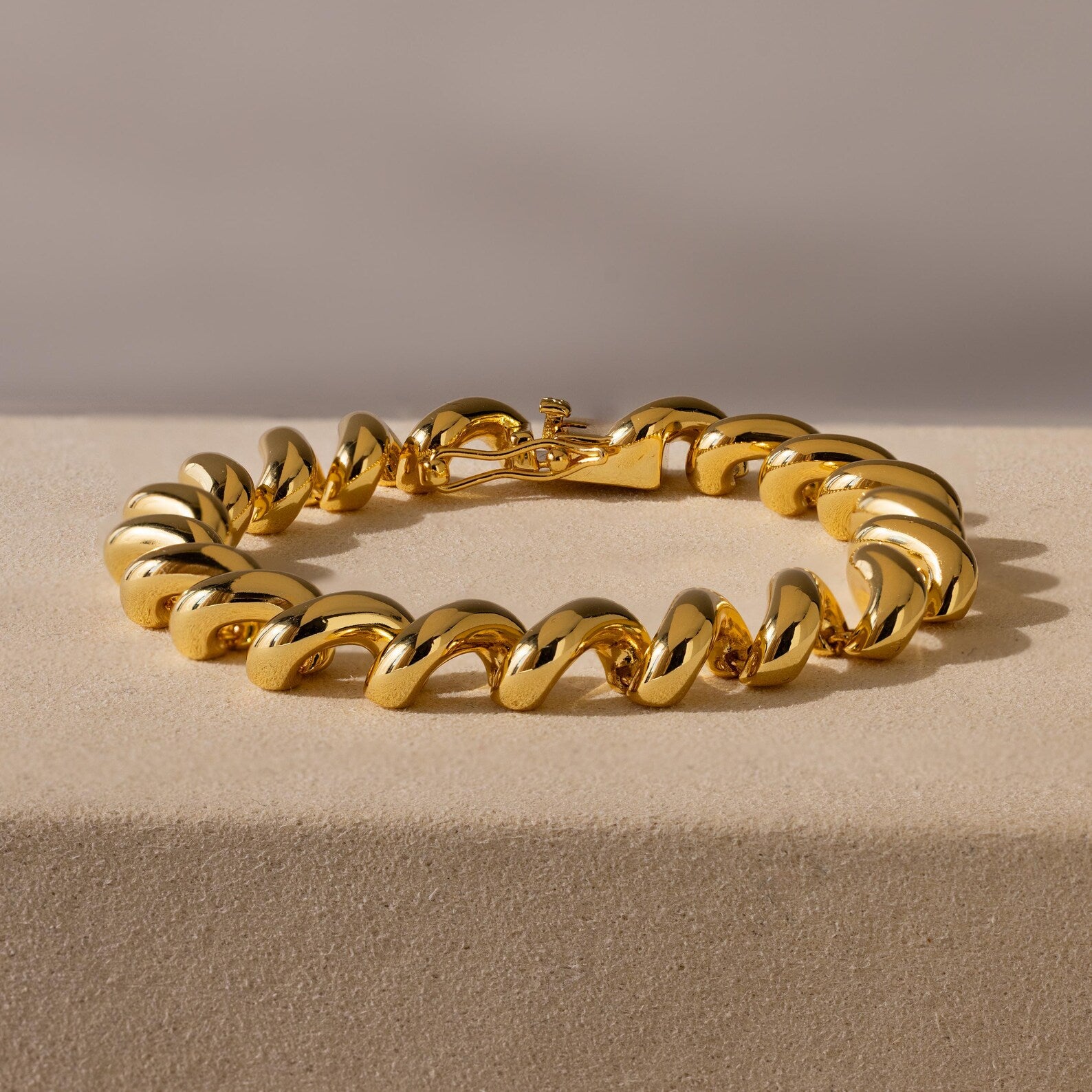 The Twisted Chain Bracelet features chunky gold, bold shining links displayed on a beige surface, radiating effortless glamour against a neutral background.