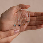 A hand holds five Custom Cut Birthstone Necklaces, each featuring a unique colored gemstone pendant—ideal as customizable, personalized jewelry for any occasion.
