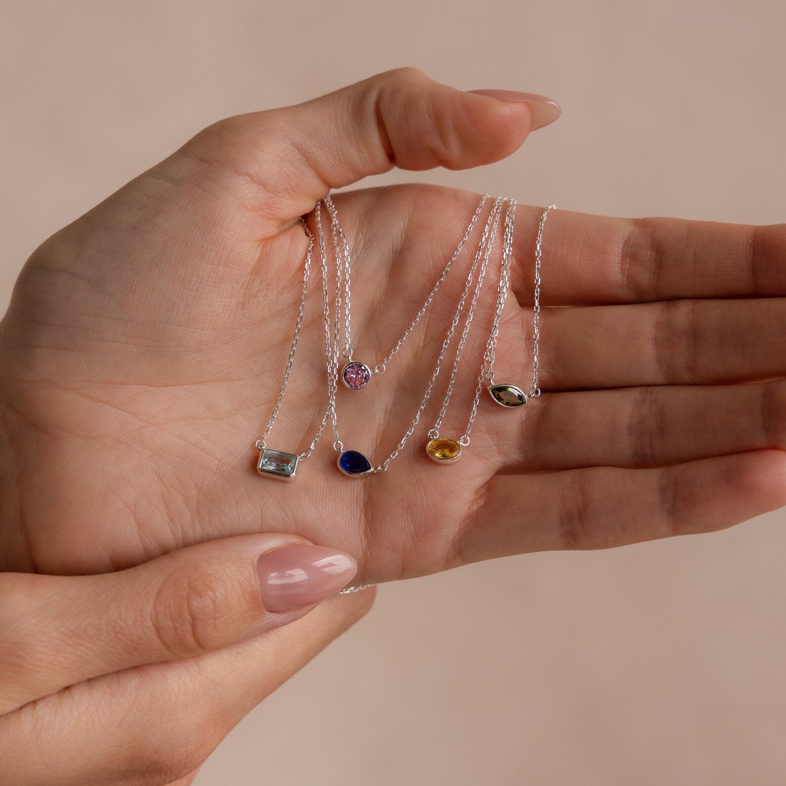 A hand holds five Custom Cut Birthstone Necklaces, each featuring a unique colored gemstone pendant—ideal as customizable, personalized jewelry for any occasion.
