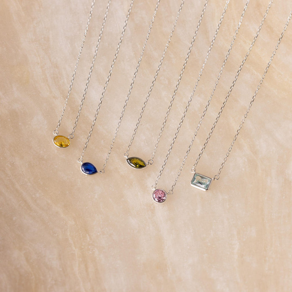 Five silver necklaces with various colored gemstone pendants, including the Custom Cut Birthstone Necklace, are displayed on a beige textured surface—perfect for those seeking personalized jewelry.
