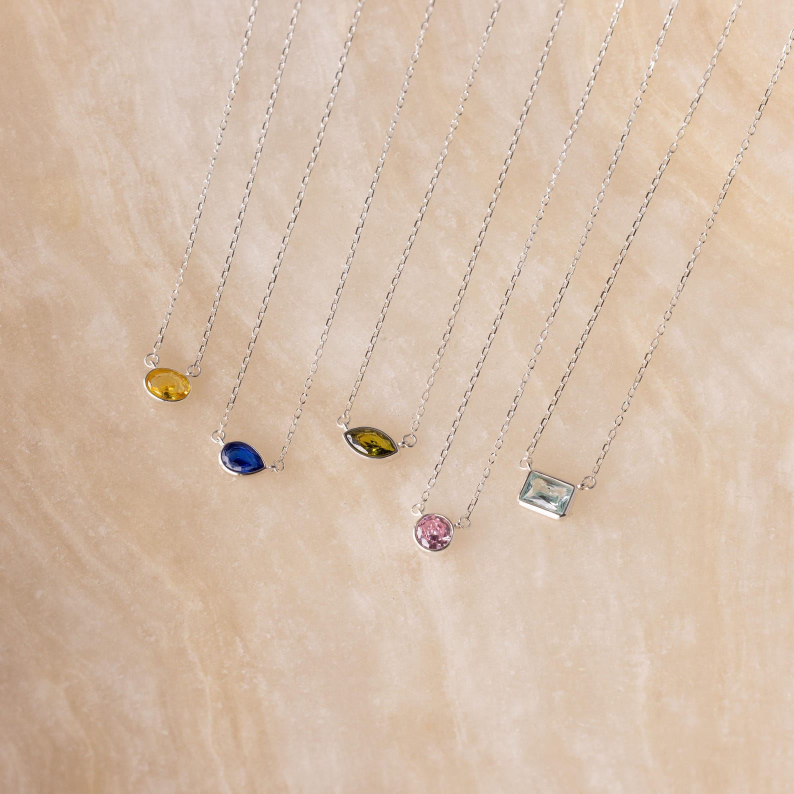 Five silver necklaces with various colored gemstone pendants, including the Custom Cut Birthstone Necklace, are displayed on a beige textured surface—perfect for those seeking personalized jewelry.