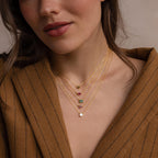 A woman wears layered gold necklaces, featuring the Custom Cut Birthstone Necklace with colorful gemstone pendants and a brown pinstripe blazer—ideal for fans of personalized jewelry.
