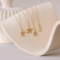 Four Large Pave Initial Necklaces with gold initial charms—K, M, R, and S—each accented with sparkling diamonds, are displayed on a curved beige surface.