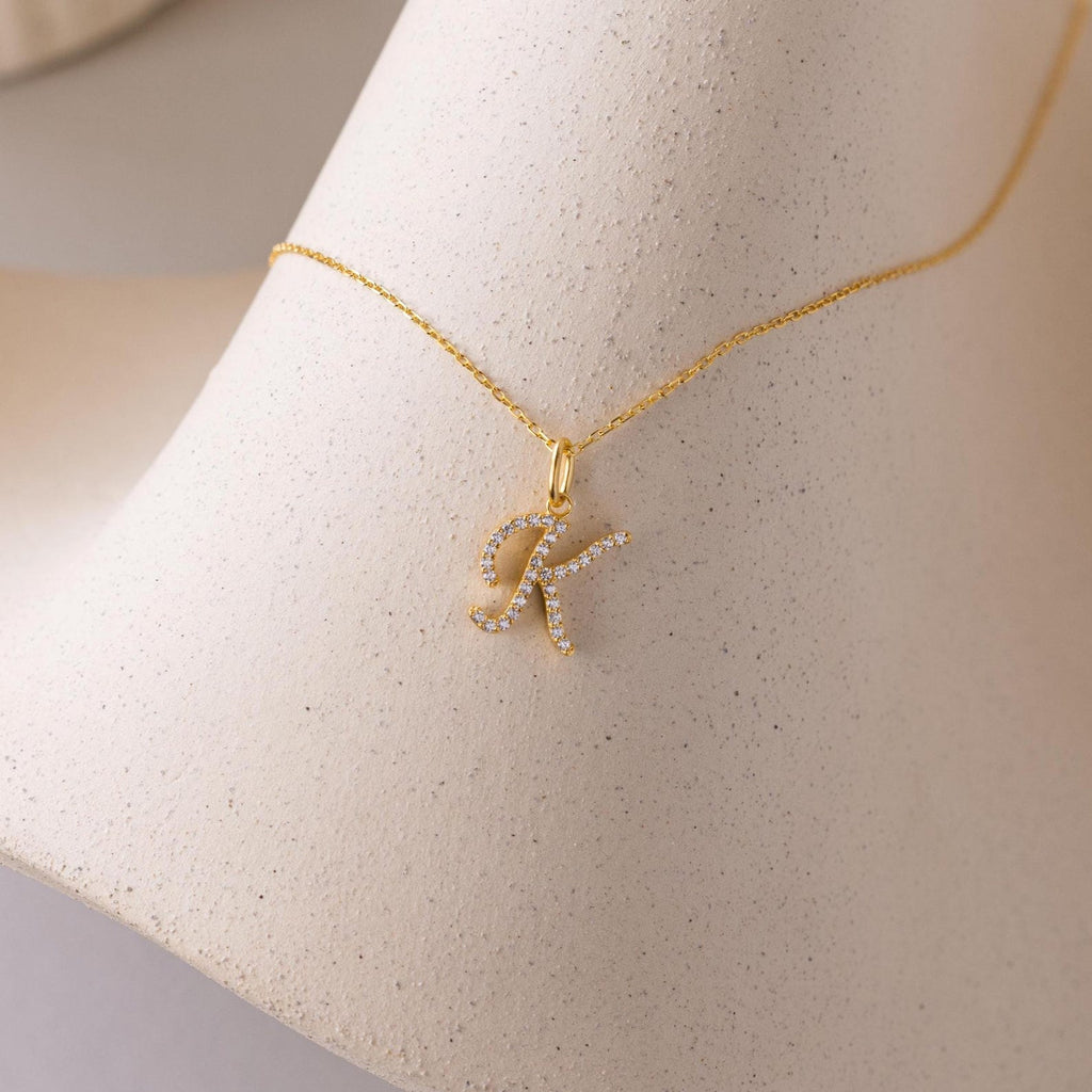 The Large Pave Initial Necklace features a jeweled letter K pendant with sparkling diamonds, displayed on a textured, light-colored surface.