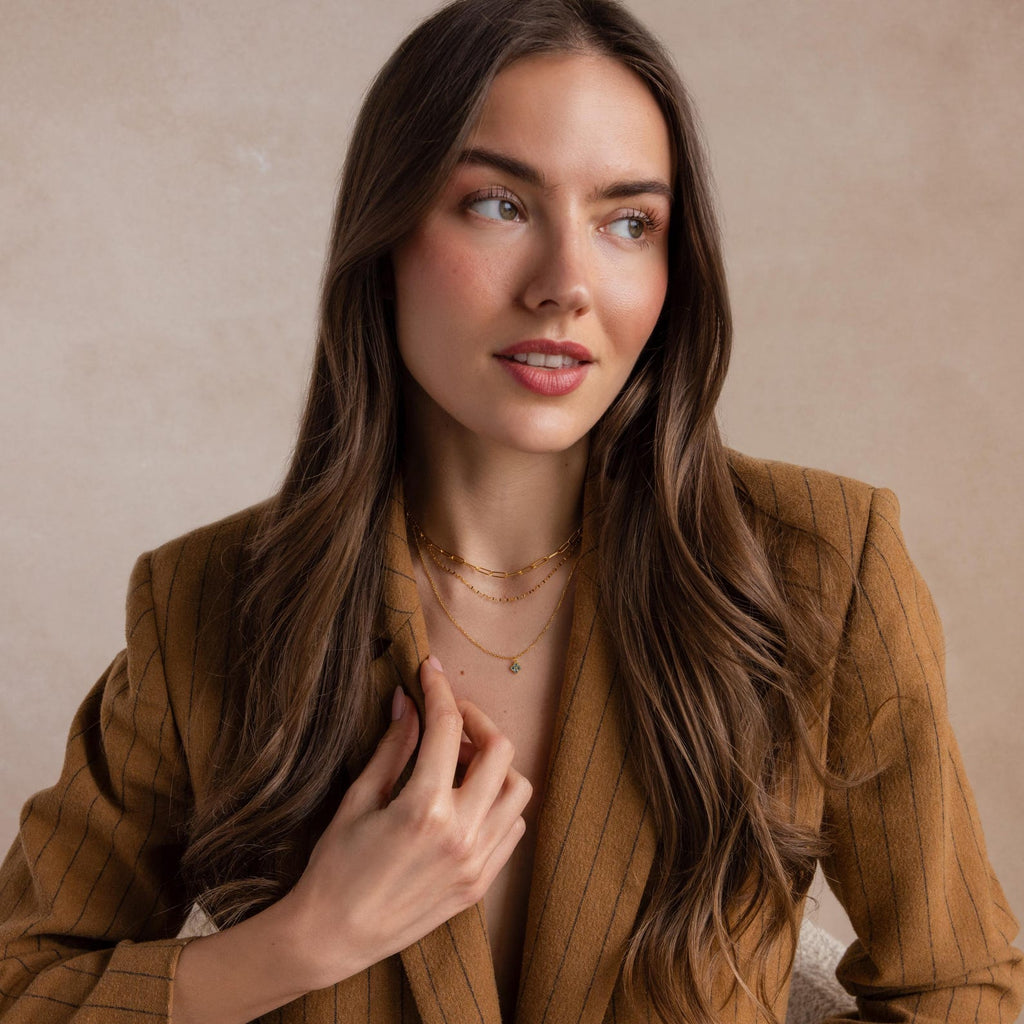 A woman with long brown hair in a brown pinstripe blazer looks away, touching her layered gold necklaces featuring the Clover Birthstone Necklace.