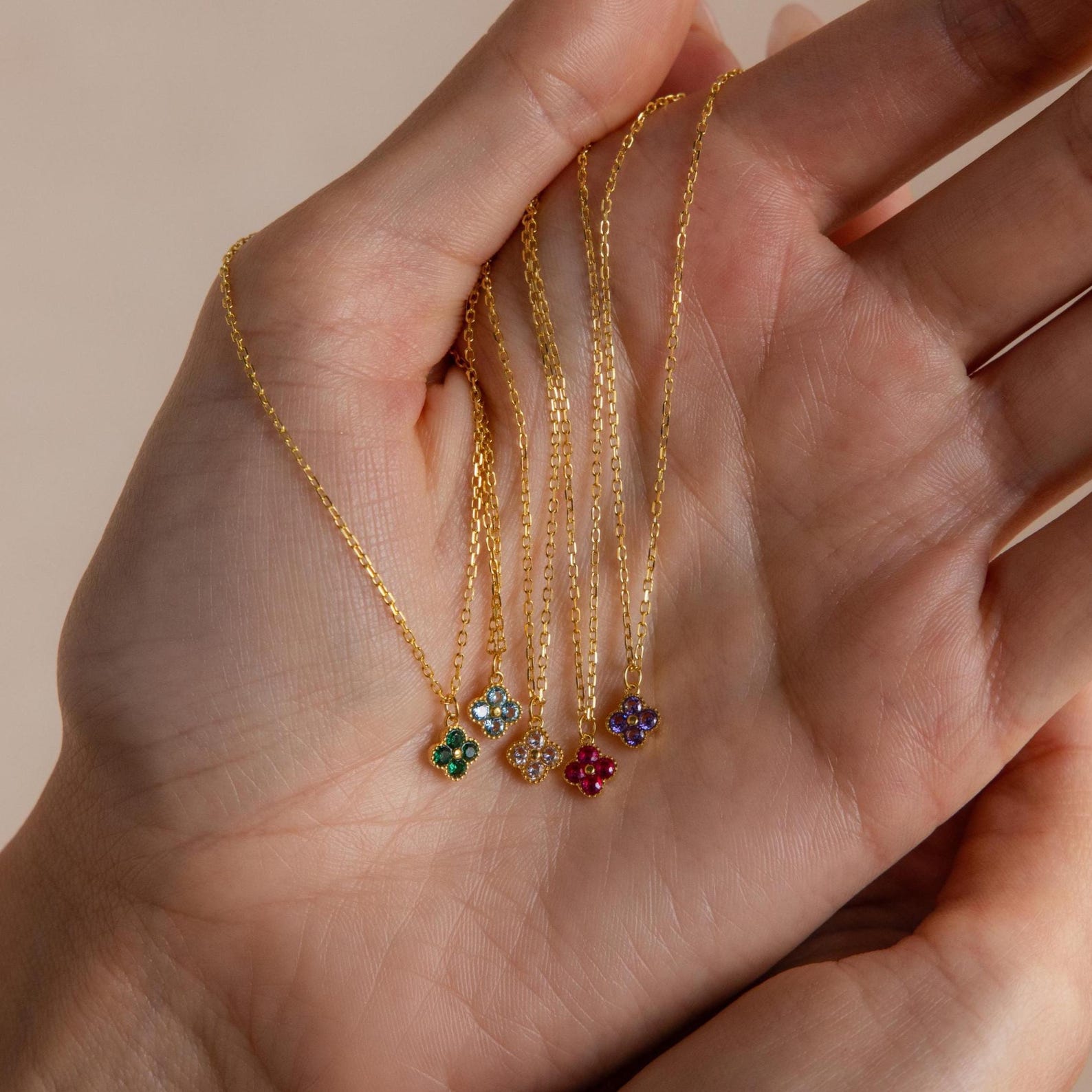 A hand holds five gold necklaces, each with a small flower-shaped pendant, including the Clover Birthstone Necklace.