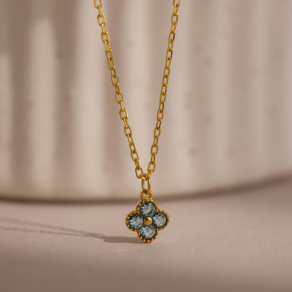 Clover Birthstone Necklace featuring a gold chain and a small blue flower-shaped pendant, photographed against a soft, neutral background.