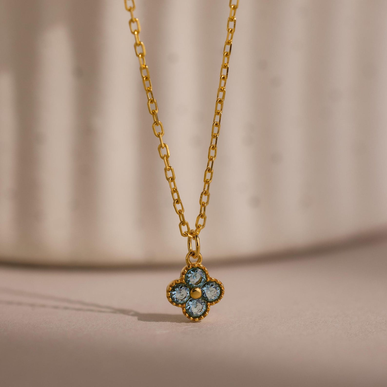 Clover Birthstone Necklace featuring a gold chain and a small blue flower-shaped pendant, photographed against a soft, neutral background.