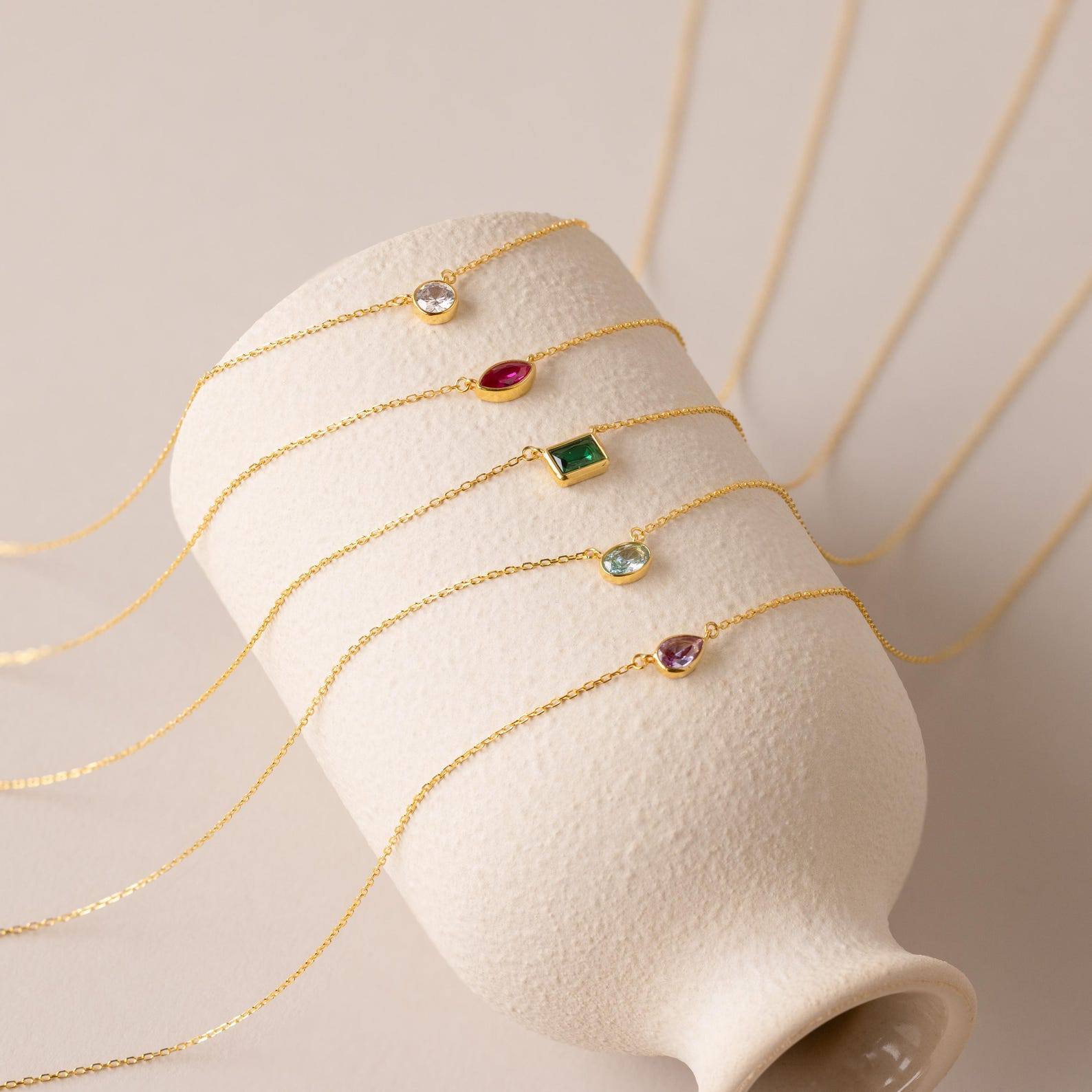 Five delicate gold chain bracelets with vibrant gemstone charms are draped over a textured ceramic vase, ideal for layering or pairing with the Custom Cut Birthstone Necklace for a personalized jewelry look.