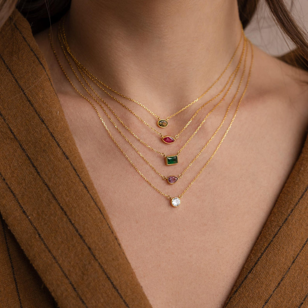A person wears a brown jacket and layered gold necklaces, including the Custom Cut Birthstone Necklace—a perfect example of personalized jewelry with customizable gemstone pendants.