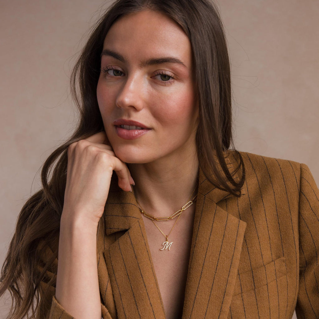 Woman in a brown pinstripe blazer, smiling softly and wearing layered gold necklaces, including the Large Pave Initial Necklace featuring sparkling diamond stones.