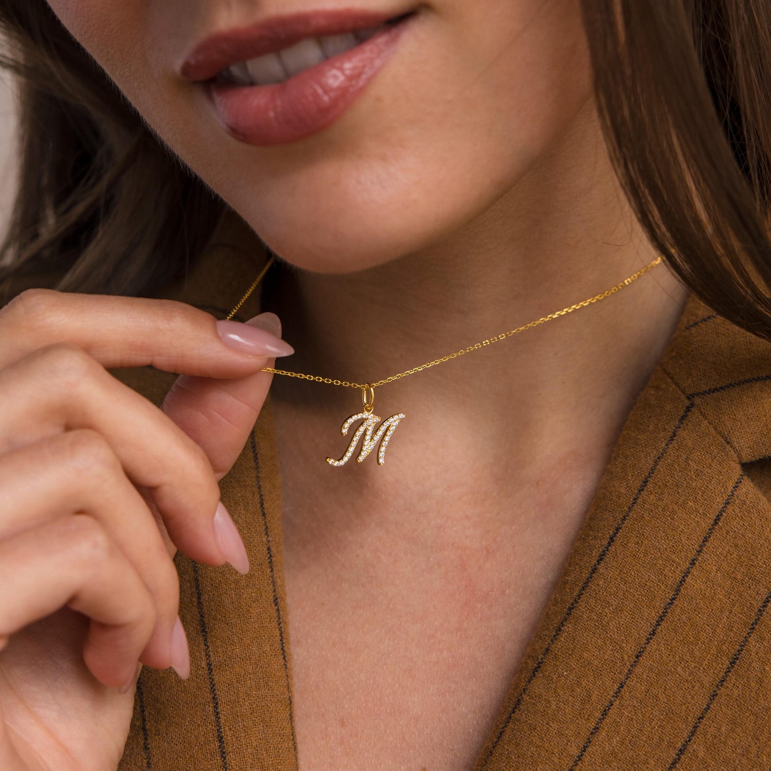 A woman in a brown blazer holds a Large Pave Initial Necklace with an M charm encrusted with diamonds near her collarbone.
