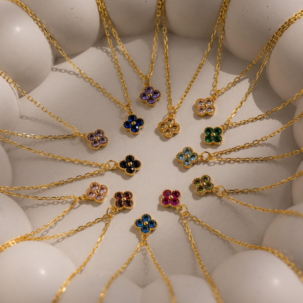 The Clover Birthstone Necklace, featuring a gold chain and colorful flower-shaped pendant, is arranged in a circular pattern with other clover charm necklaces on a white surface.