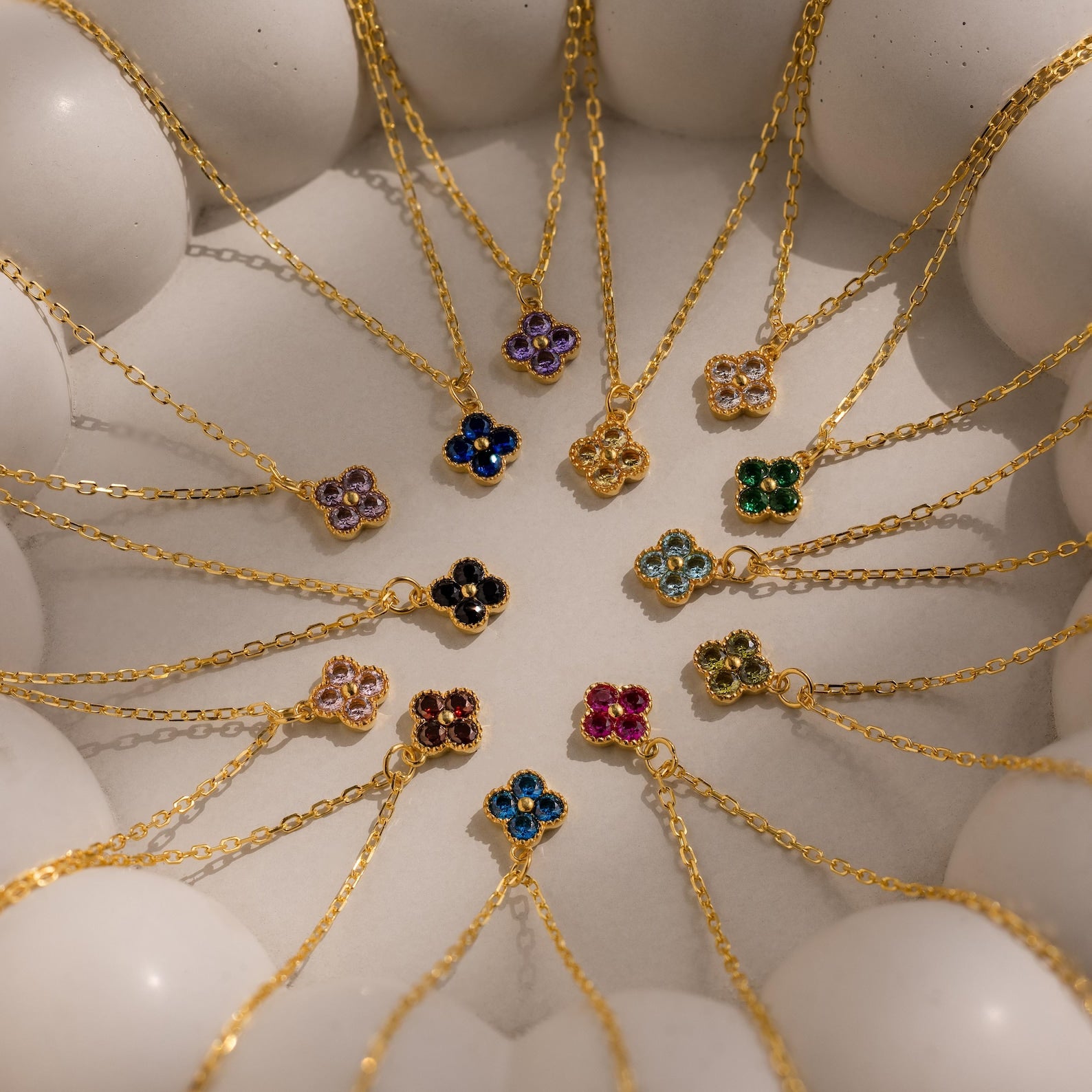 The Clover Birthstone Necklace, featuring a gold chain and colorful flower-shaped pendant, is arranged in a circular pattern with other clover charm necklaces on a white surface.