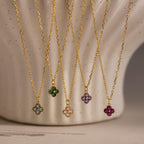 Five gold necklaces with colorful gemstone flower pendants, including the Clover Birthstone Necklace, are displayed hanging against a textured cream background.