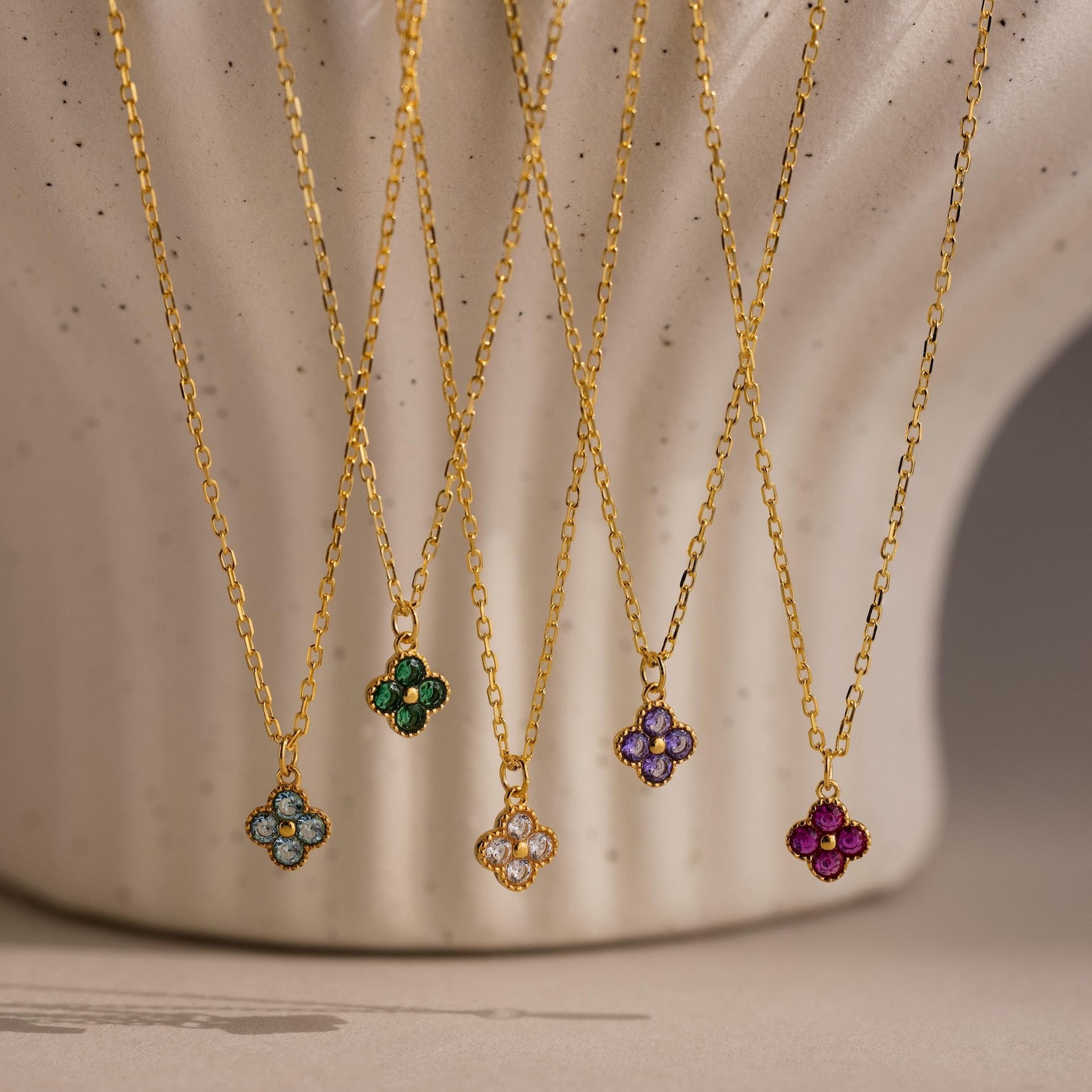 Five gold necklaces with colorful gemstone flower pendants, including the Clover Birthstone Necklace, are displayed hanging against a textured cream background.