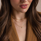 A woman wears the Clover Birthstone Necklace, featuring a four-leaf clover charm, layered with gold chains and paired with a brown pinstripe blazer. The image is shown from shoulders to lips.