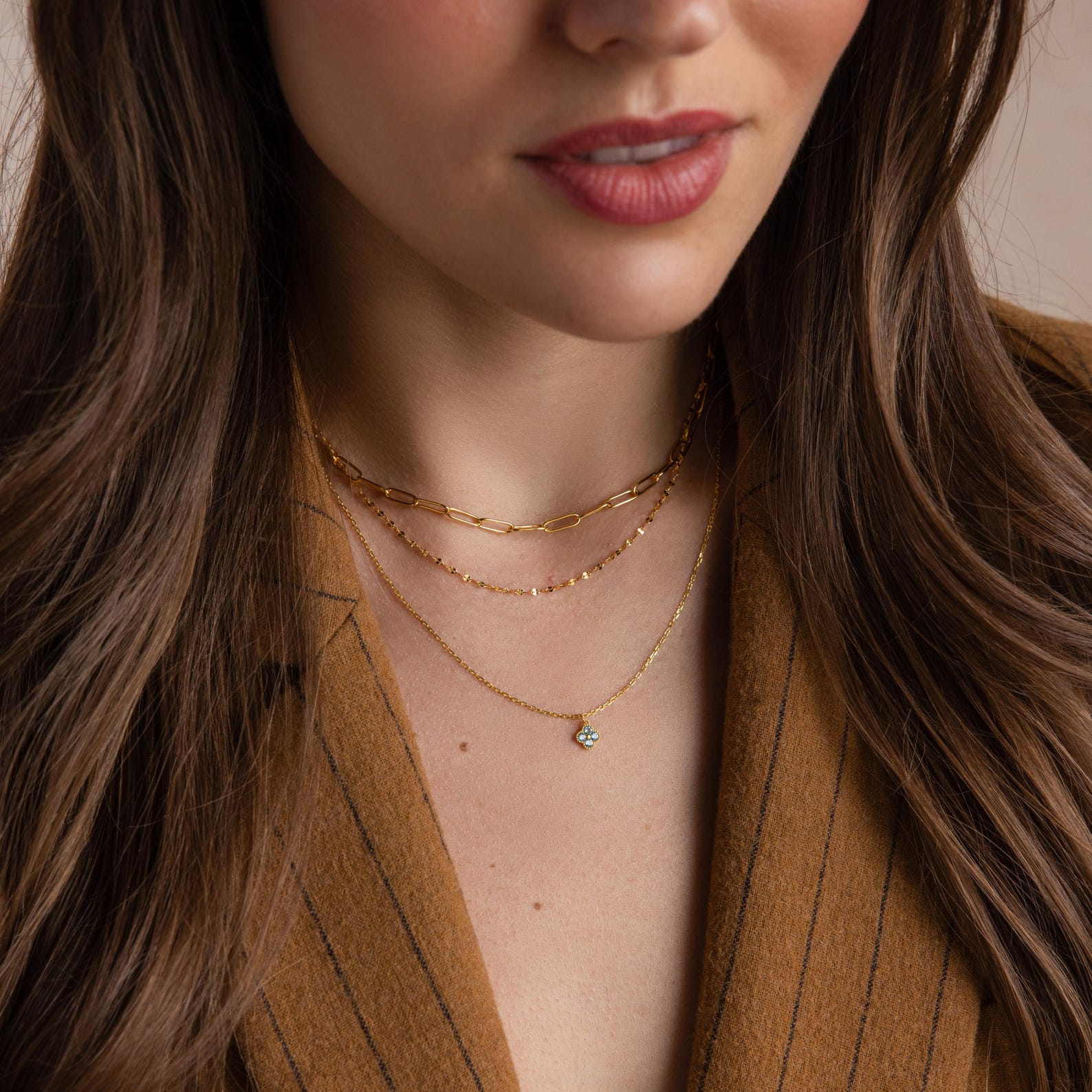 A woman wears the Clover Birthstone Necklace, featuring a four-leaf clover charm, layered with gold chains and paired with a brown pinstripe blazer. The image is shown from shoulders to lips.
