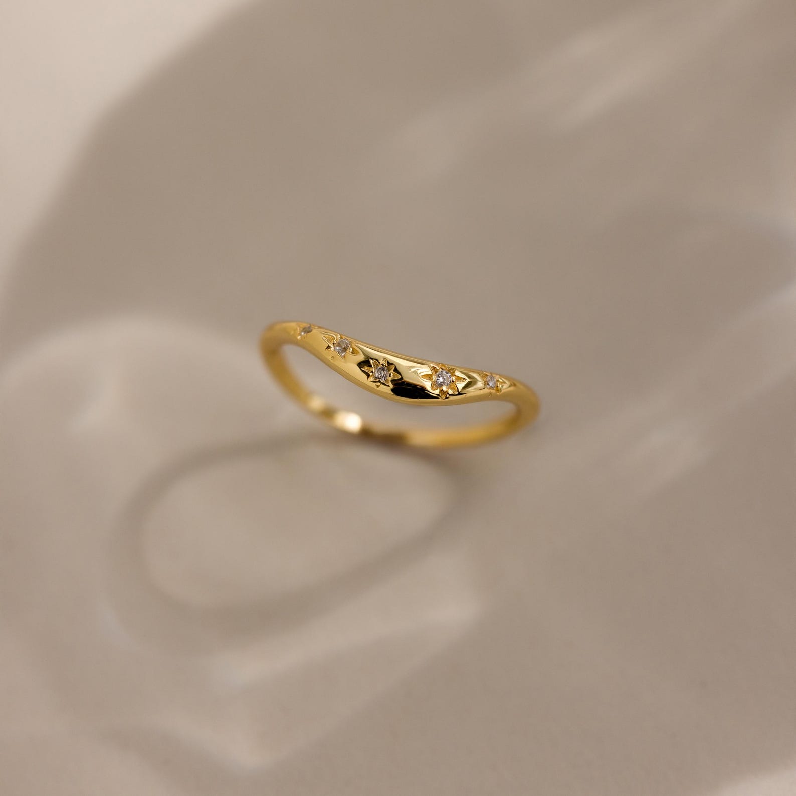 The North Star Curve Dome Ring features diamond star accents and delicate star-shaped engravings, with gemstones shimmering as it casts a soft shadow on a beige surface.