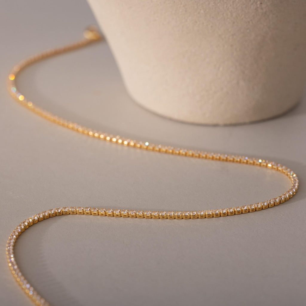 The Dainty Diamond Tennis Necklace & Bracelet Set in 18K Gold, pictured beside a ceramic pot on a beige surface, offers a refined and elegant option for anniversary jewelry.