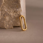 An 18k gold oval carabiner clasp from the Multipurpose Mixed Metal Clasp Set rests against a textured beige stone on a neutral background, ideal for stacking rings or expanding your jewelry collection.