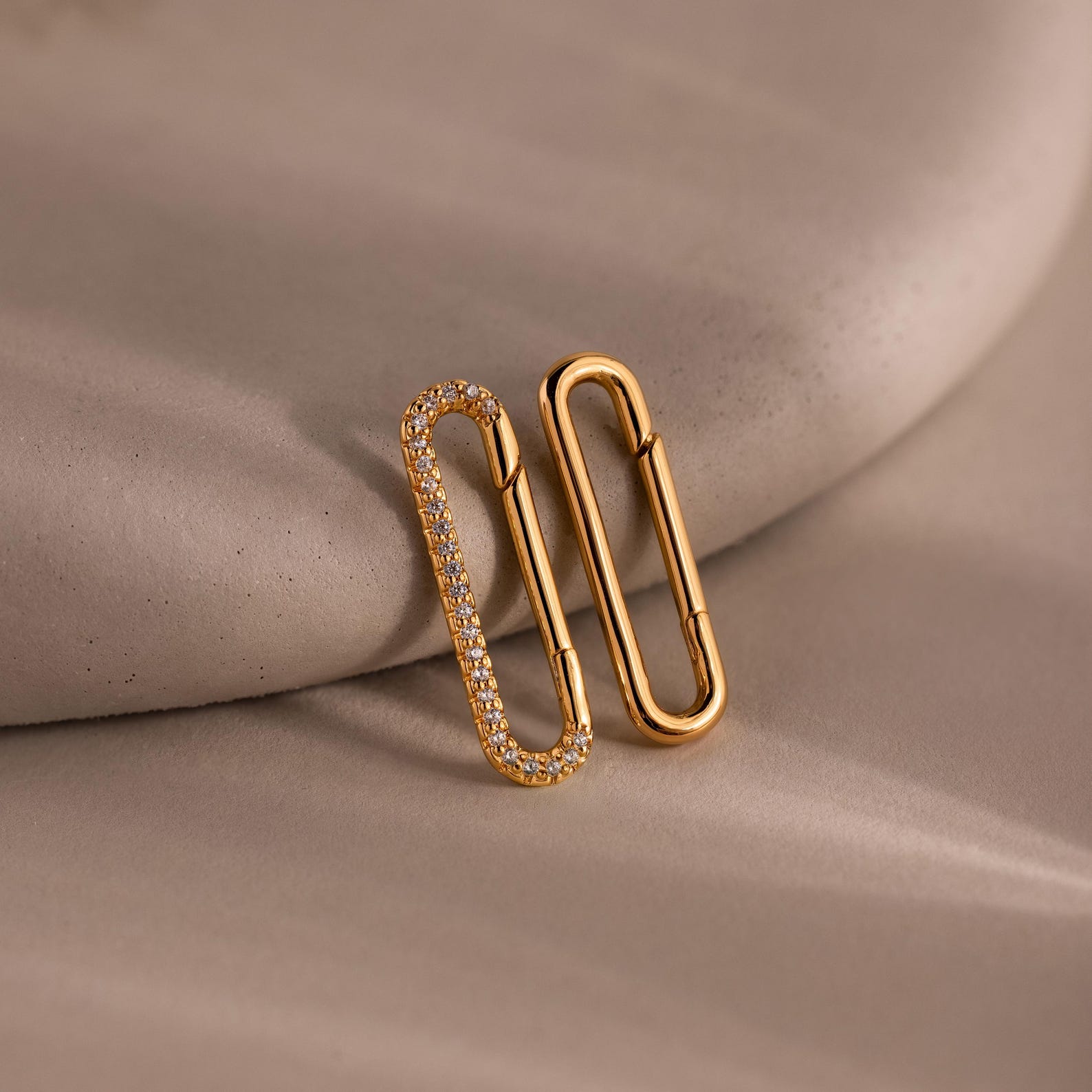 The Multipurpose Pave Link Clasp Set in 18K Gold features two paperclip-shaped earrings—one with sparkling stones—displayed on a soft beige surface, offering versatile styling options.