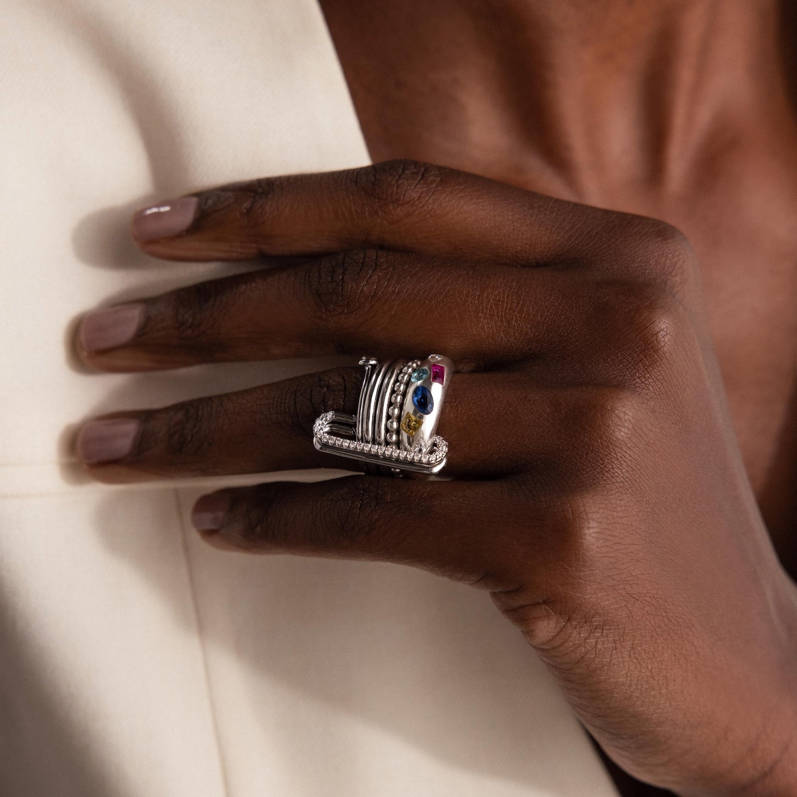 A manicured hand displays a modern silver multi-band ring with colorful gemstones, styled next to a chic 18k white gold Multipurpose Pave Link Clasp Set for a contemporary jewelry look.