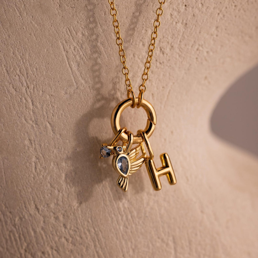 The Hummingbird Charm in 18K Gold features a sparkling diamond accent, bird and letter H charms, shown against a beige textured background—an elegant and meaningful gift for someone special.