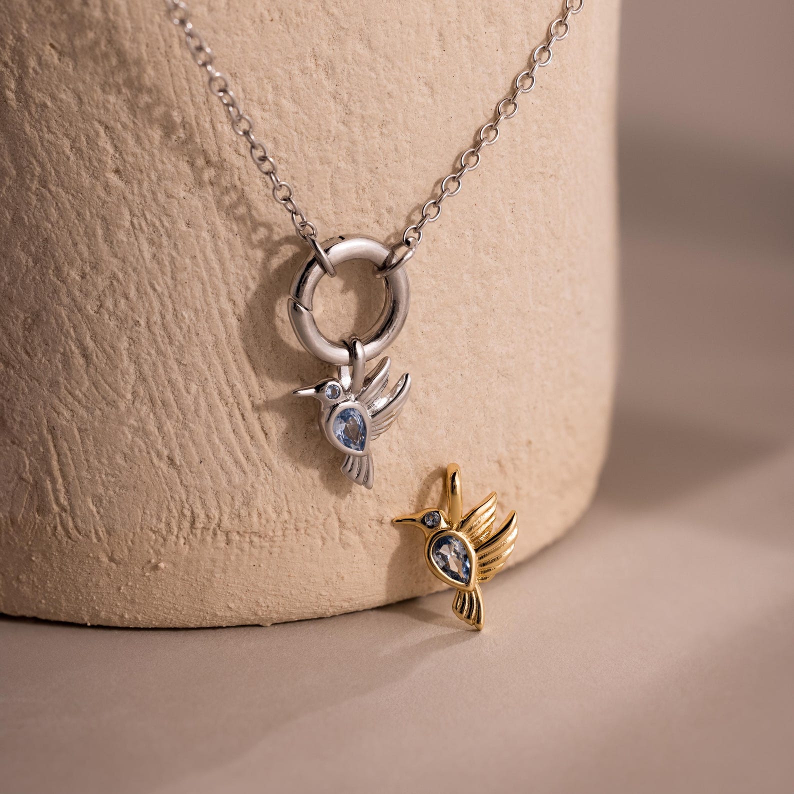 Silver and gold Hummingbird Charm pendants adorned with blue gemstones—one on a chain, one displayed on a textured surface—make a thoughtful gift for any occasion.