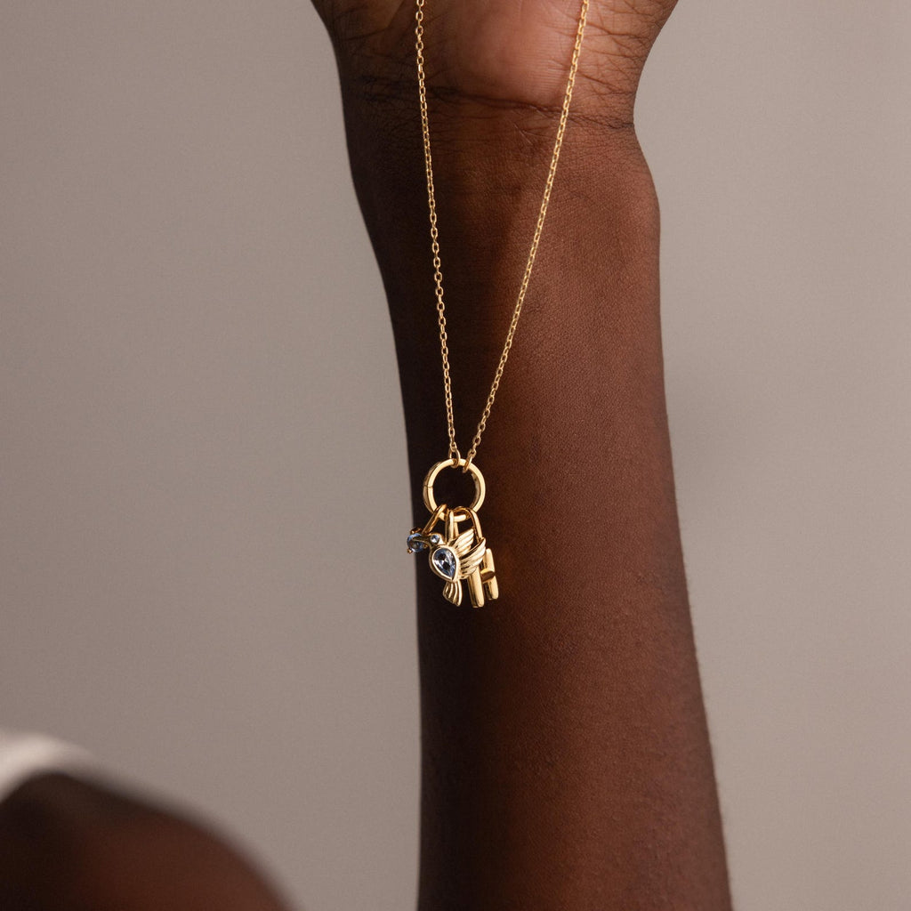 The Hummingbird Charm gold chain necklace, featuring delicate gold charms and a pear-cut diamond, is held by a raised hand against a neutral background, making it a meaningful gift.