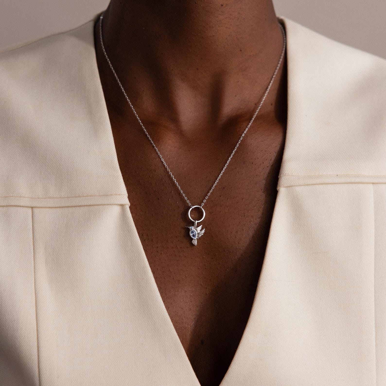 A silver necklace featuring the Hummingbird Charm with pear-cut diamond detail, worn over a cream V-neck blazer.