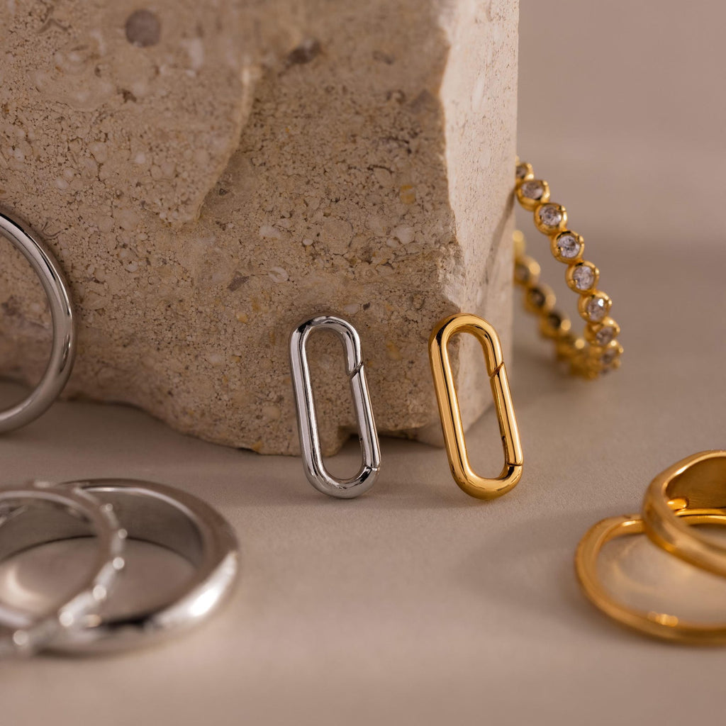 Two metal earrings—one silver, one 18k gold—stand upright against a textured stone, with rings nearby, perfect for stacking. Elevate your style with the Multipurpose Mixed Metal Clasp Set for elegant versatility.