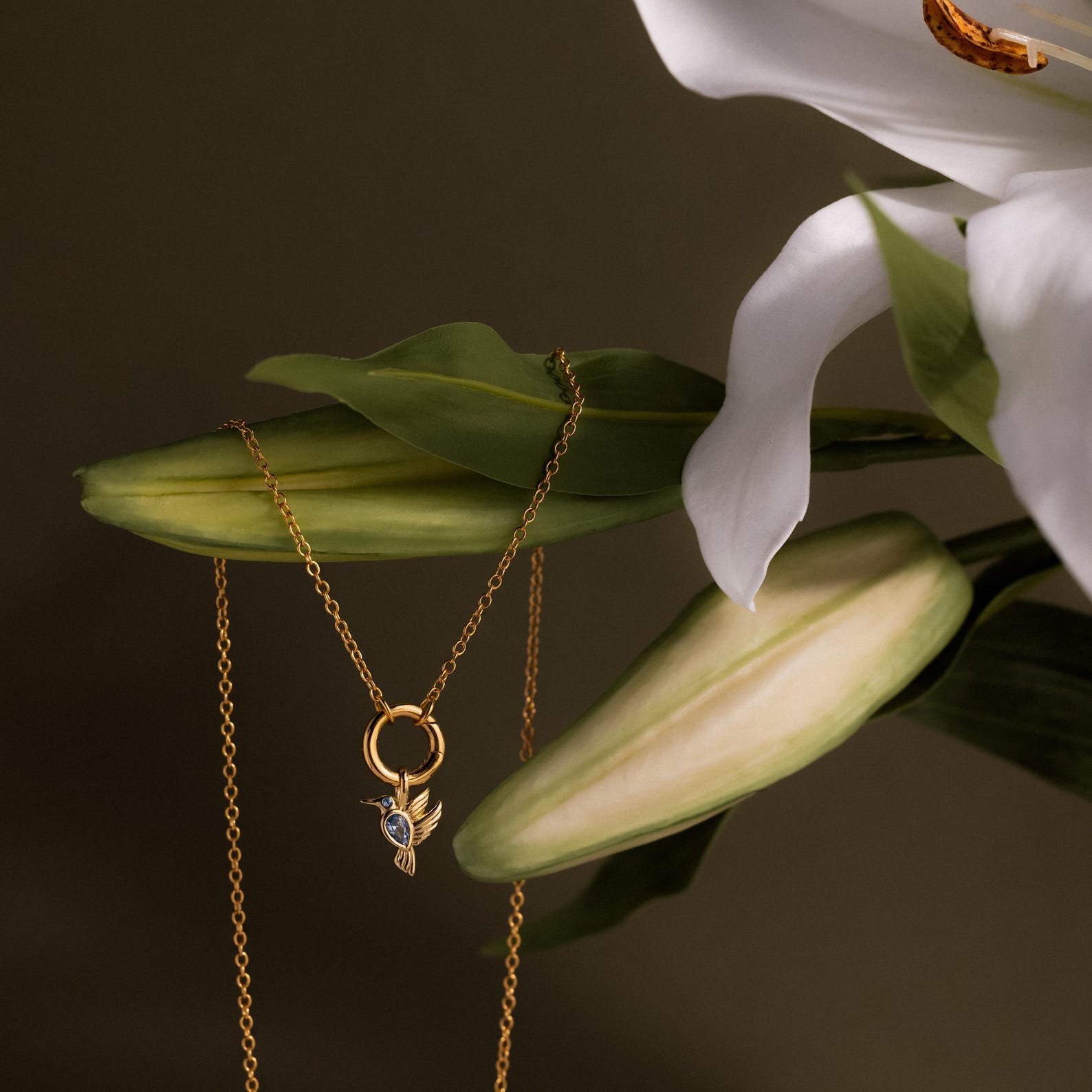 The Hummingbird Charm in 18K Gold pendant necklace draped on green leaves and white lilies in soft light—a meaningful gift for any occasion.