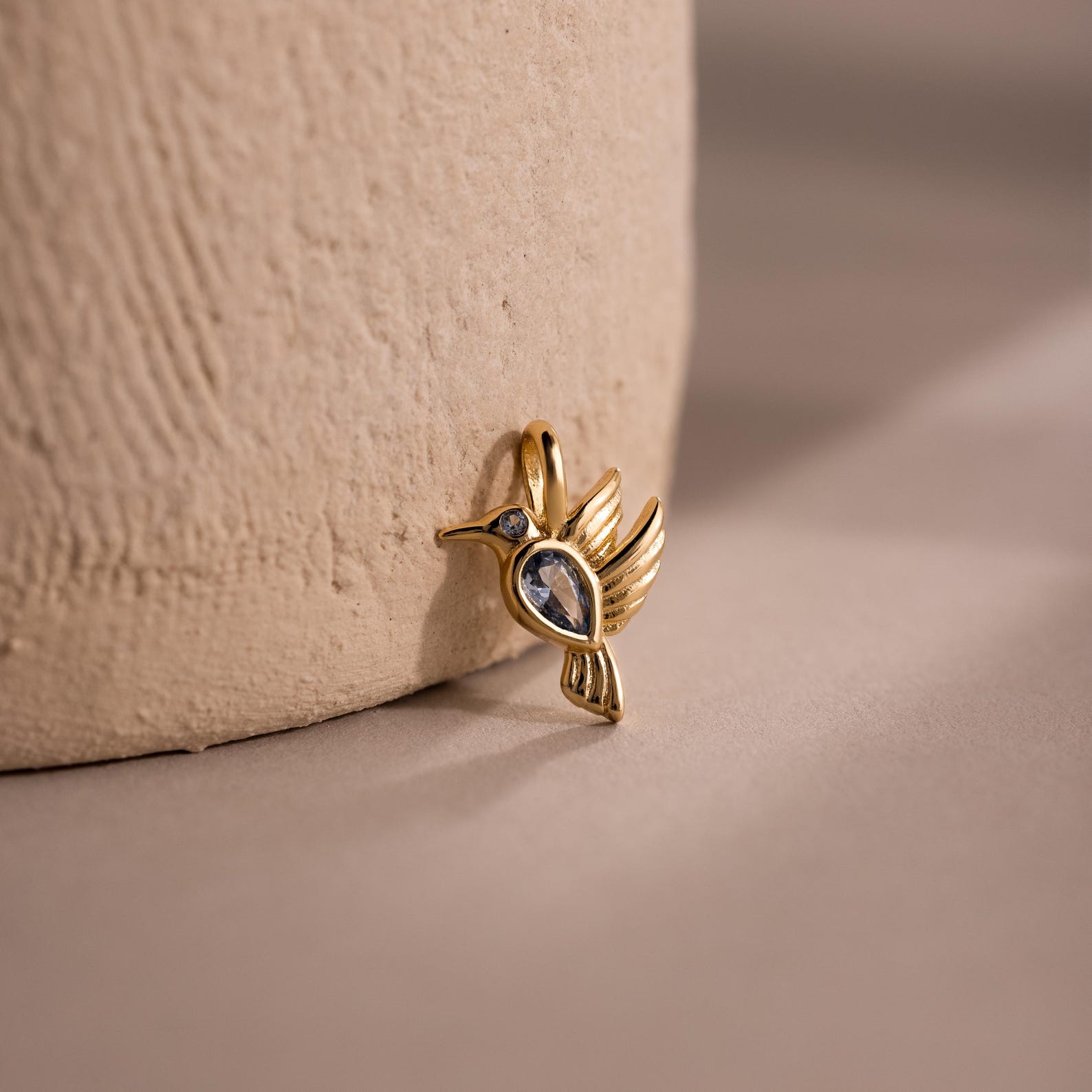 The Hummingbird Charm in 18K Gold features a clear gemstone body and diamond accent, displayed on a textured beige surface—a thoughtful gift for someone special.