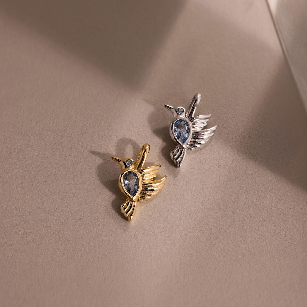 Two Hummingbird Charm pendants, one gold and one silver with a blue gemstone center, rest on a beige surface—a perfect meaningful gift for someone special.
